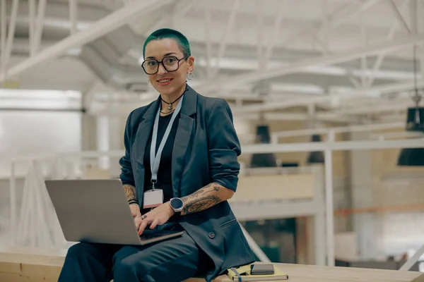 Smiling tattooed female freelancer with green short hair working on laptop while sitting in office