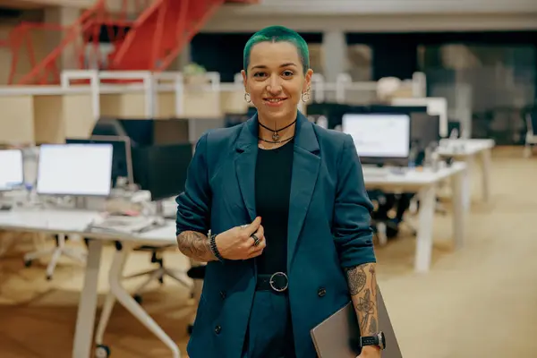 Smiling tattooed business woman with green short hair holding laptop standing in modern office