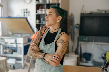 Dizayn stüdyosundaki baskı makinesinin arka planında elleri çapraz duran bir kadın işçi.