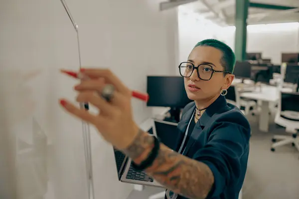 Stylish tattooed business woman working on laptop and making notes on flipchart on office background