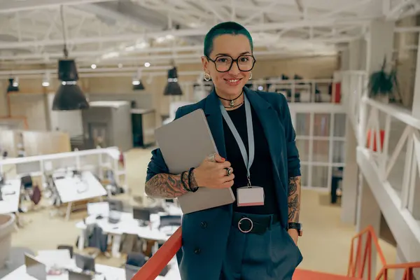 Creative tattooed female freelancer in glasses with laptop standing on modern coworking background
