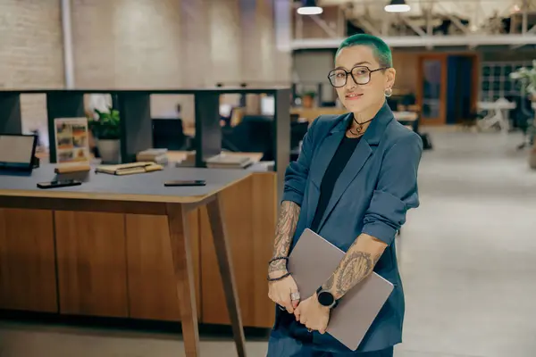 Creative tattooed female freelancer with laptop standing on modern coworking background