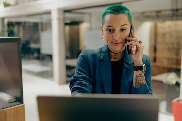 Pretty tattooed female business owner is use laptop while talking phone sitting in coworking