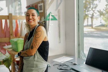 Tasarım stüdyosunda dizüstü bilgisayarı olan önlüklü bir kadın çalışan kameraya bakıyor.