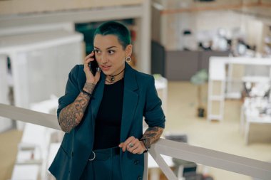 Yaratıcı dövmeli iş kadını, modern ofiste müşterisiyle cep telefonundan konuşuyor.