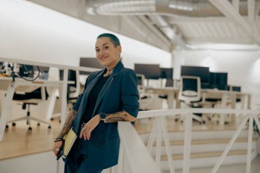 Gülümseyen, dövmeli, yeşil kısa saçlı iş kadını elinde dizüstü bilgisayarla modern ofiste duruyor.