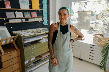 Gülümseyen kadın tasarımcı atölyenin arka planında duruyor ve kameraya bakıyor.