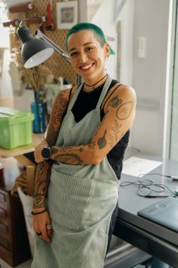 Gülümseyen kadın tasarımcı kendi atölyesinin arka planında oturuyor ve kameraya bakıyor 