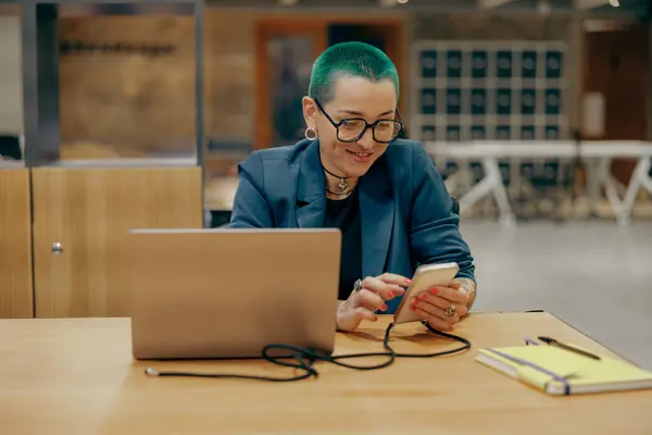 Pretty tattooed female business owner is use laptop while use phone sitting in coworking