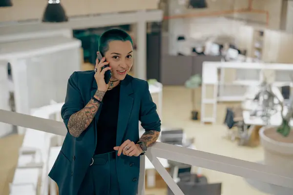 Pretty tattooed female business owner is talking mobile phone while standing in modern coworking