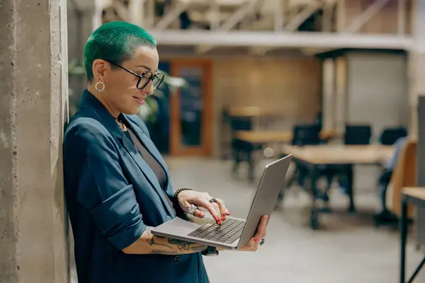 Creative tattooed business woman with green short hair working on laptop while standing in office