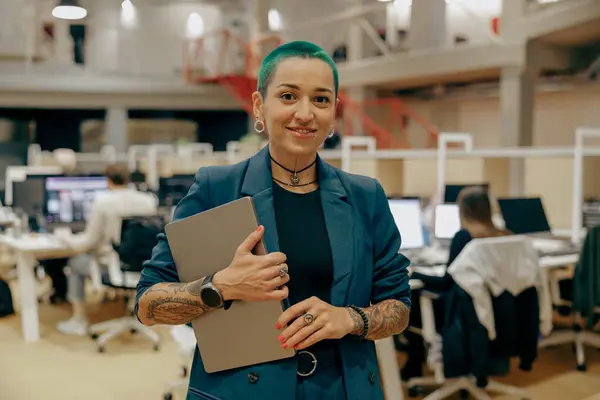 Smiling tattooed business woman with green short hair holding laptop standing in modern office