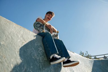 Mavi gökyüzüne karşı kaykaylı rampada oturan rahat giyimli genç erkek patencinin alçak açısı.