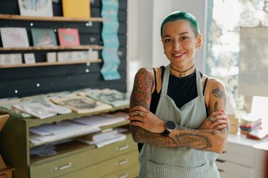 Pozitif kadın işçi dizayn stüdyosunun arka planında çapraz ellerle duruyor ve kameraya bakıyor.