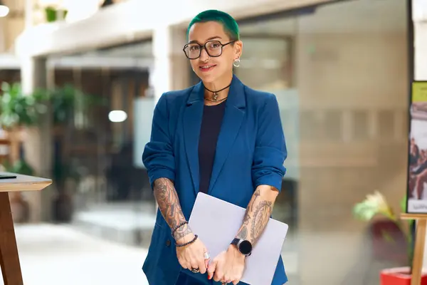 A woman with electric blue hair, tattoos, and glasses, wearing a denim blazer and formal attire, is smiling while holding a laptop at an event
