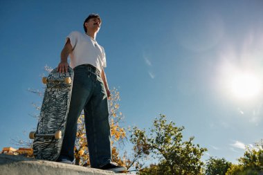 Güneşli bir günde kaykay parkında elinde kaykayla duran mutlu erkek kaykaycının alçak açısı.