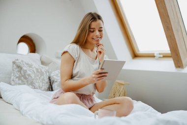 Bir kadın ahşap bir yatakta oturuyor ve not defterine notlar yazıyor. 