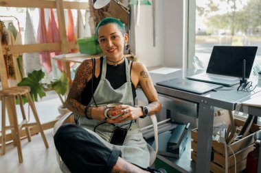 Yaratıcı kadın işçi kendi tasarım stüdyosunun arka planında oturuyor ve kameraya bakıyor