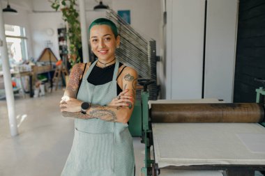 Gülümseyen kadın tasarımcı atölyenin arka planında ellerini çapraz yapıp kameraya bakıyor.