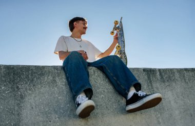 Mavi gökyüzüne karşı kaykaylı rampada oturan rahat giyimli genç erkek patencinin alçak açısı.