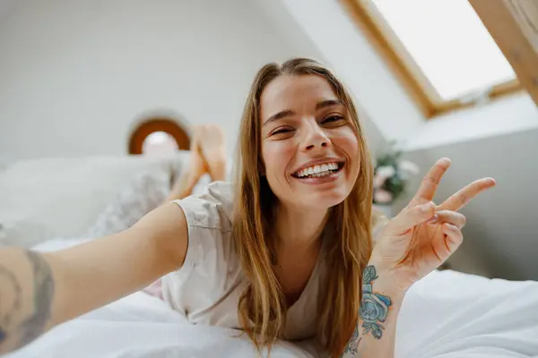 Happy woman is lying on a bed, flashing a peace sign gesture with her fingers while taking a selfie