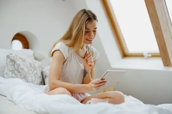 Young smiling woman making notes in note pad while sitting on bed at home