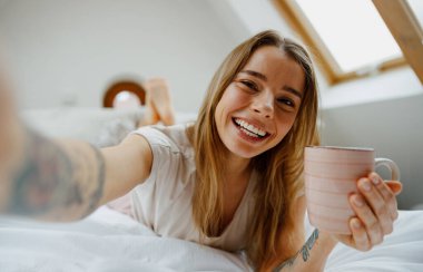 Kadın elinde bir fincan kahve tutarken gülümsüyor ve selfie çekmek için kolunu kaldırıyor.