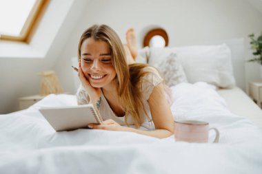 Pijamalı gülümseyen kadın tahta bir yatakta oturmuş not defterindeki notları okuyor.
