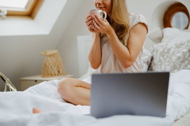 Bir kadın yatağa oturur, elinde kahveyle dizüstü bilgisayar kullanır. 