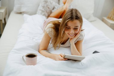 Evdeki yatakta oturmuş not defterinde notlar okuyan gülümseyen genç bir kadın. 