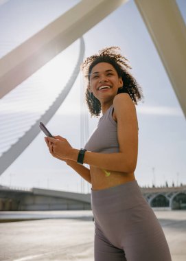 Spor kıyafetleriyle gülümseyen kadın sporcu eğitimden sonra dışarıda duruyor ve cep telefonu kullanıyor.