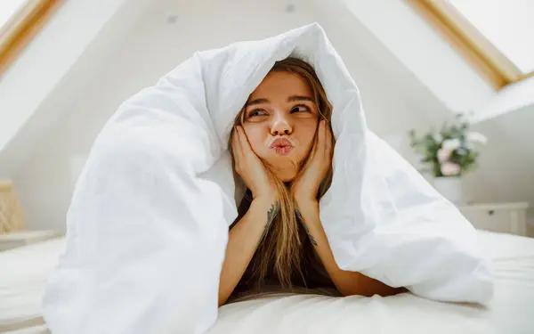 A woman is laying under a blanket on a bed, making a funny face in a gesture of leisure