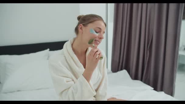 バスローブの女性は 彼女の顔にローラーを使用してベッドに座っています 魅惑的なジェスチャーと楽しいイベント — ストック動画