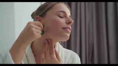 Yeşim bir silindir ve gua sha taşıyla yüz masajı yapan bir kadını izleyin. Burun, yanak, dudak ve daha fazlası için teknikler gösterirken gülümsüyor. Güzellik tavsiyeleri ve rahatlama yöntemleri sunuyor.