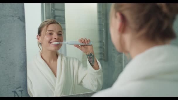 女性は鏡の前に立って笑顔で彼女の手で歯を磨いている 彼女は彼女の目の近くにしわをスポットし 彼女の反射にそれを指摘する — ストック動画