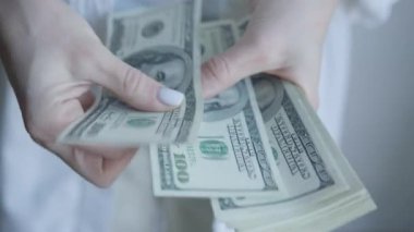 Bir kadının elleri, zenginlik, finans ve manuel para işleme kavramlarını vurgulayan, kalın bir yığın 100 dolarlık banknotları sayarken görülüyor.