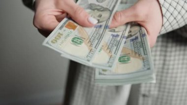 Detaylı bir çekim, ekose takım elbiseli bir kadını titizlikle 100 dolarlık banknotları sayarken yakalıyor ve zenginlik ve finans kavramını vurguluyor.