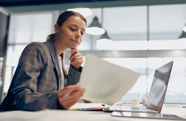Belgelerle çalışan gülümseyen iş kadını ofiste otururken dizüstü bilgisayar kullanıyor. 
