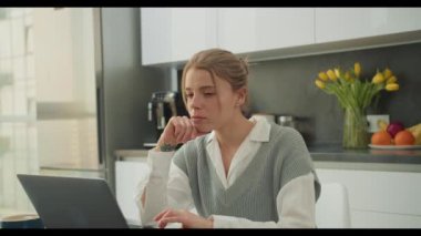 Bir kadın, modern bir mutfakta çalışırken yoğun bir şekilde laptopuna odaklanır. Sahne, uzak bir çalışma ortamında konsantrasyon ve verimliliği vurguluyor.