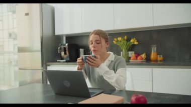 Modern bir mutfakta dizüstü bilgisayarı üzerinde çalışırken bir kadın kahve molasından zevk alır. Sahne, rahat bir ev ortamında üretkenlik ve rahatlama arasındaki dengeyi vurguluyor..