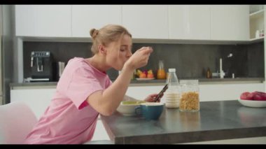 Pembe gömlekli genç bir kadın modern bir mutfakta telefonunu karıştırırken sağlıklı bir kahvaltıda mısır gevreği ve süt yiyor. Görüntü teknolojinin ve sağlıklı beslenmenin karışımını yakalıyor.