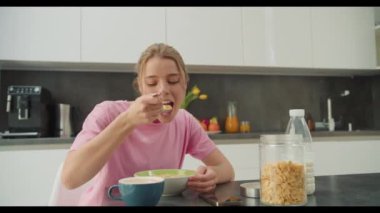 Pembe gömlekli genç bir kadın modern bir mutfakta sağlıklı bir kahvaltıda mısır gevreği ve süt yiyor. Görüntü besleyici bir sabah rutininin ve rahat bir yaşam tarzının özünü yakalıyor.