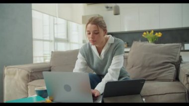 Parlak bir oturma odasında rahat bir kanepede otururken dikkatle dizüstü bilgisayarını ve tabletini kullanan genç bir kadın. Sahne, modern ev yaşamını ön plana çıkarıyor.