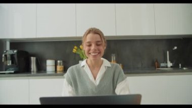Genç bir kadın, video görüşmesi sırasında neşeli bir şekilde el sallıyor. Parlak ve modern bir mutfakta oturuyor. Görüntü, çağdaş bir ev ortamında dost canlısı ve uzak iletişimi yakalıyor..