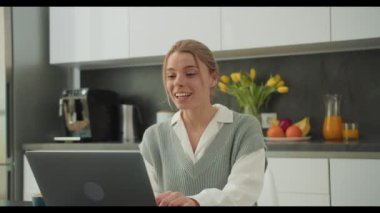 Modern bir mutfakta dizüstü bilgisayarıyla video görüşmesi yapan bir kadın sıcak bir şekilde gülümsüyor. Bu görüntü, çağdaş bir evde sanal iletişimin ve uzaktan çalışmanın özünü yakalar.