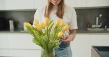 Modern bir mutfakta sarı lalelere hayran genç bir kadın, sakince çiçeklere doğru eğiliyor, dingin bir ifade yaratıyor genç sarışın bir kadın nazikçe bir buket çiçeğe doğru eğiliyor.