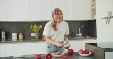 Genç bir kadın taze bir salata hazırlıyor, domates ve yapraklı yeşilliklerle dolu bir kaseye mozzarella topları ekliyor. Etrafı dolgun çan biberi ve elma gibi parlak meyvelerle çevrili.