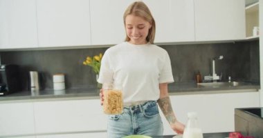 Genç bir kadının parlak, modern bir mutfakta mısır gevreğine taze süt dökerken çekilmiş yakın plan bir fotoğrafı. Sahne, besleyici bir kahvaltının basitliğini ve tazeliğini canlı detaylarla yakalıyor.