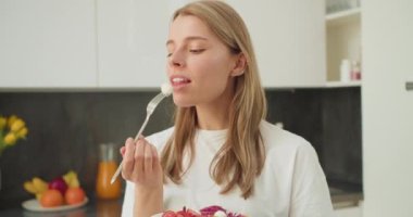 Genç bir kadın, modern bir mutfakta taze mozzarella ısırığının tadını çıkararak sağlıklı beslenmenin keyfini çıkarıyor. Minimalist mutfak ve doğal ışıklandırma sahnenin basitliğini artırır