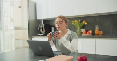Sıcak mutfak ortamında bir kadın laptopuyla uğraşırken bir fincan kahvenin tadını çıkarıyor. Sahne, ev ortamında rahatlama ve verimliliğin bir karışımını yakalıyor..